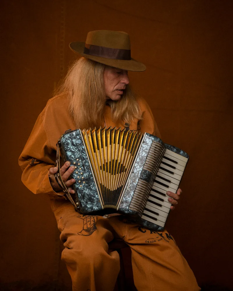 Sanfoneiro de chapéu e macacão da coleção Brasil Rural, tocando sentado no barracão